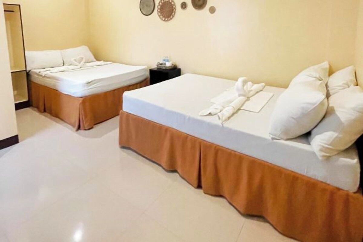 A hotel room with two beds, each with a white mattress and brown bed skirt. There is a small table with a lamp and a clock on the wall.