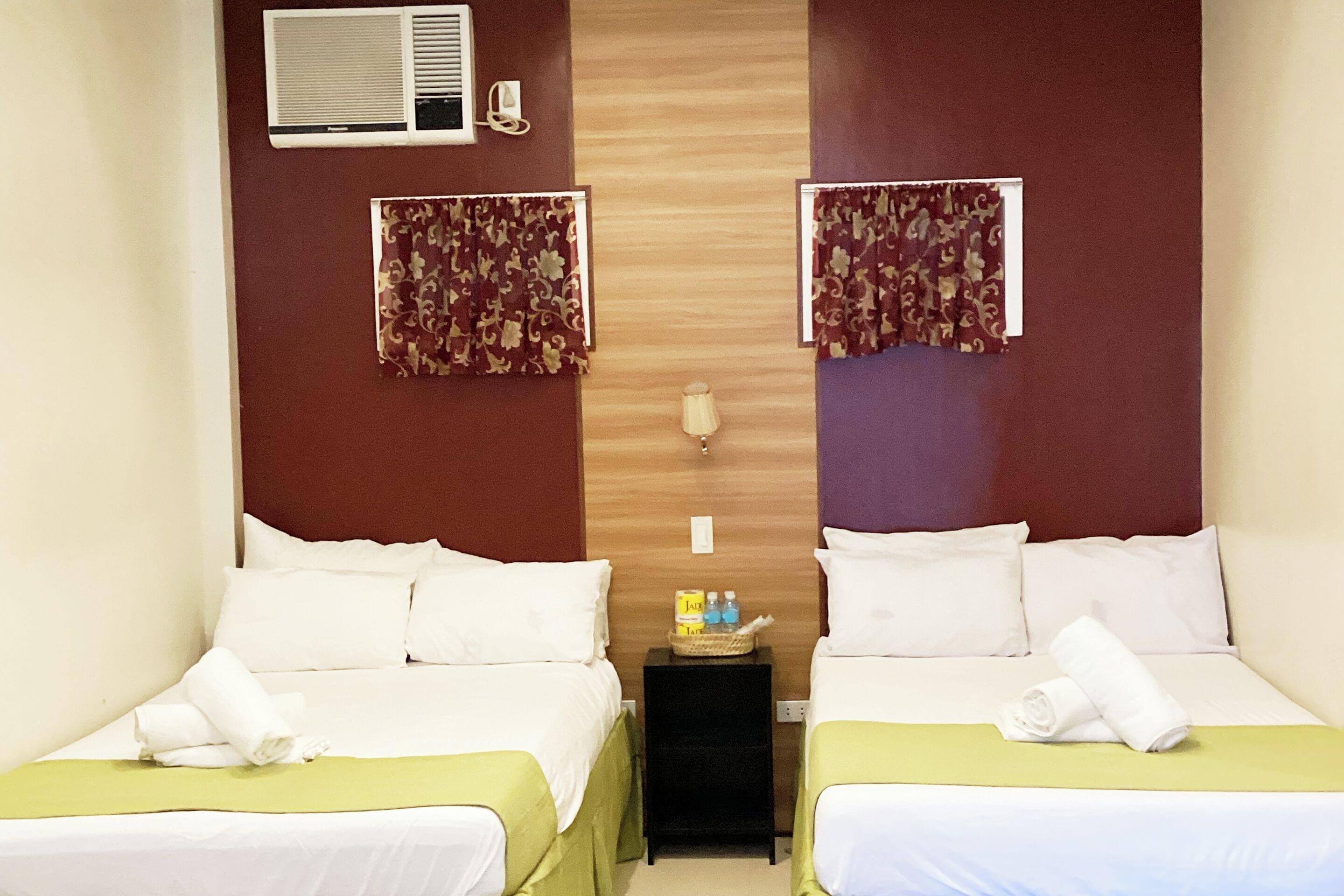 A hotel room with two beds, each with white linens and a green bedspread. There is a small table between the beds with a lamp and a basket containing toiletries.