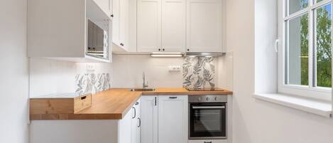 A compact kitchen with white cabinetry, a wooden countertop, a built-in oven, and a window with a view of trees.