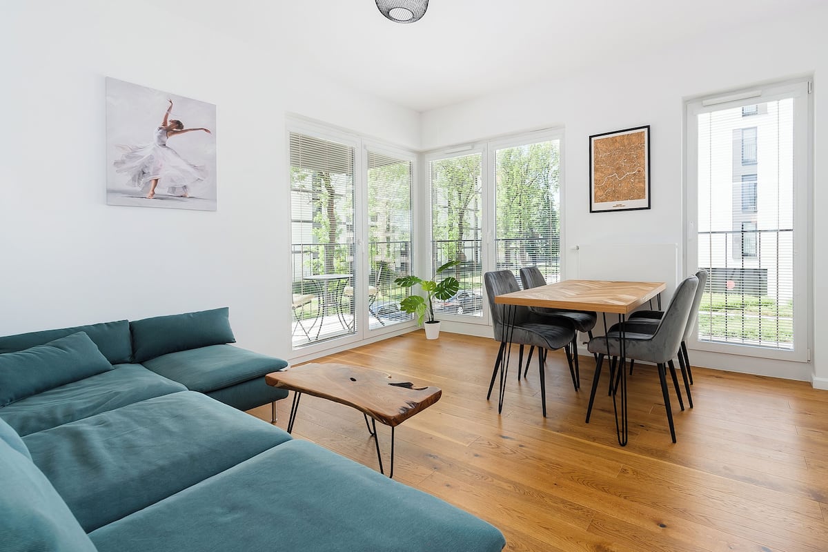 A modern living room with a teal sofa, a wooden dining table with chairs, and large windows offering a view of the outdoors.