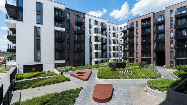 Ein modernes Apartmenthaus mit mehreren Balkonen, einem begrünten Innenhof mit Pflanzkästen und einer zentralen Holzbank.