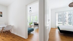 Ein modernes, helles Zimmer mit Holzboden, einem Bett, einem Schreibtisch mit Stühlen und einem Balkon mit Blick auf Grünflächen.