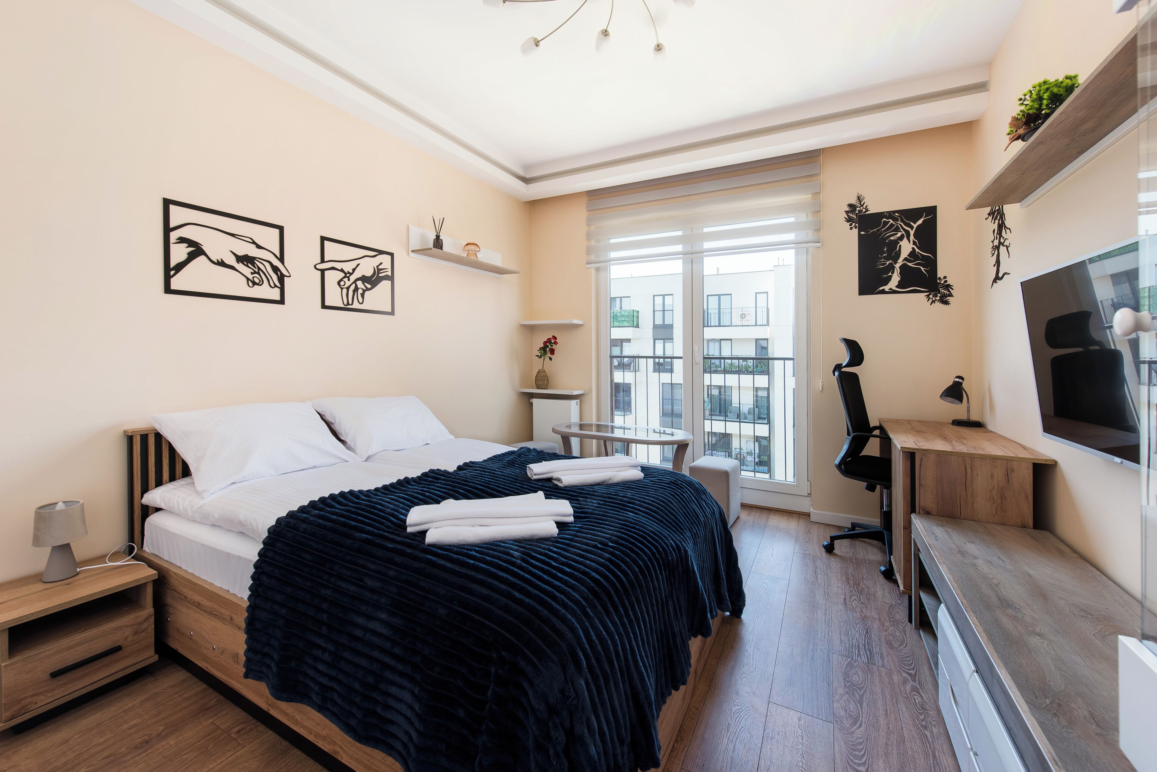 A bedroom with a bed, bedside table, lamp, TV, desk, chair, and a window with a view of buildings.