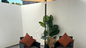 A patio area with two wicker chairs, each with a brown cushion, under a large beige umbrella. A potted plant with broad leaves is placed between the chairs. - Youradress (Casablanca)