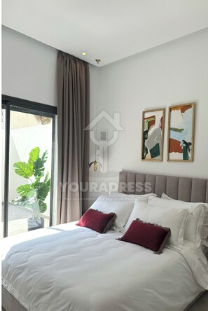 Une chambre avec un grand lit, une fenêtre avec des rideaux, une plante en pot et deux œuvres d’art encadrées accrochées au mur.