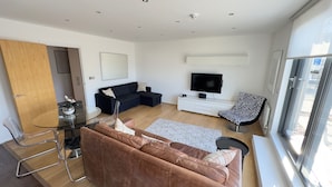 A modern living room with a brown leather sofa, a glass dining table, a black and white patterned chair, a flat-screen TV, and a large window. - Oval House Retreat in Camden Town (London)