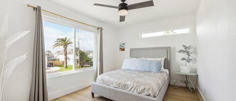 A bedroom with a bed, a ceiling fan, a window with curtains, a small table with a plant, and a view of palm trees outside.