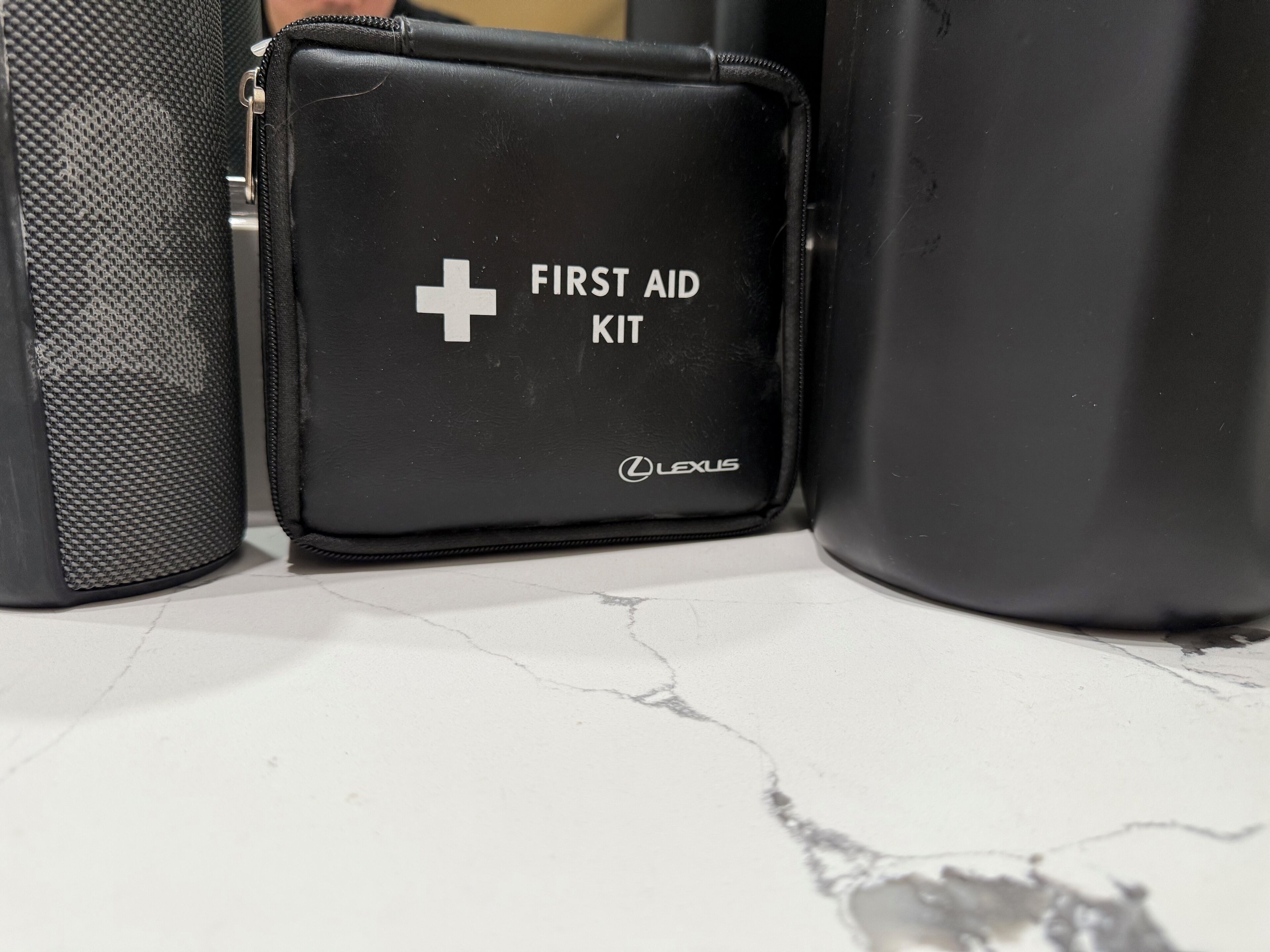 Ein schwarzes Erste-Hilfe-Set mit einem weißen Kreuz und der Aufschrift "FIRST AID", platziert auf einem Marmorboden neben einer schwarzen Tasche und einem zylindrischen Gegenstand.