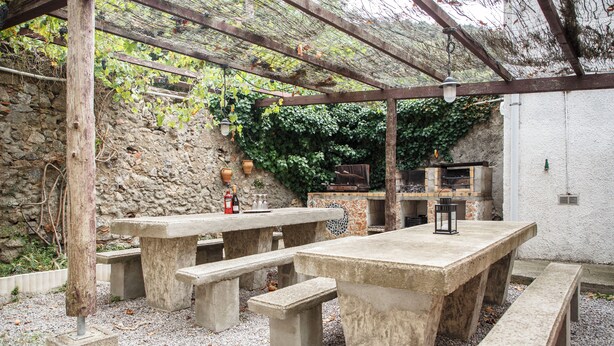 Un patio al aire libre con mesas y bancos de piedra, una pérgola y un muro de piedra con vegetación.