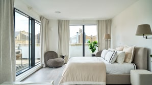 Une chambre moderne avec un grand lit, un canapé, un fauteuil et une vue sur la ville grâce à de grandes fenêtres.