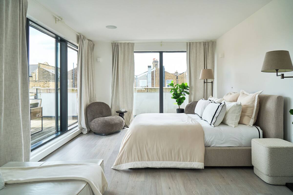 Ein modernes Schlafzimmer mit einem großen Bett, einer Couch, einem Sessel und Blick auf die Stadt durch große Fenster.