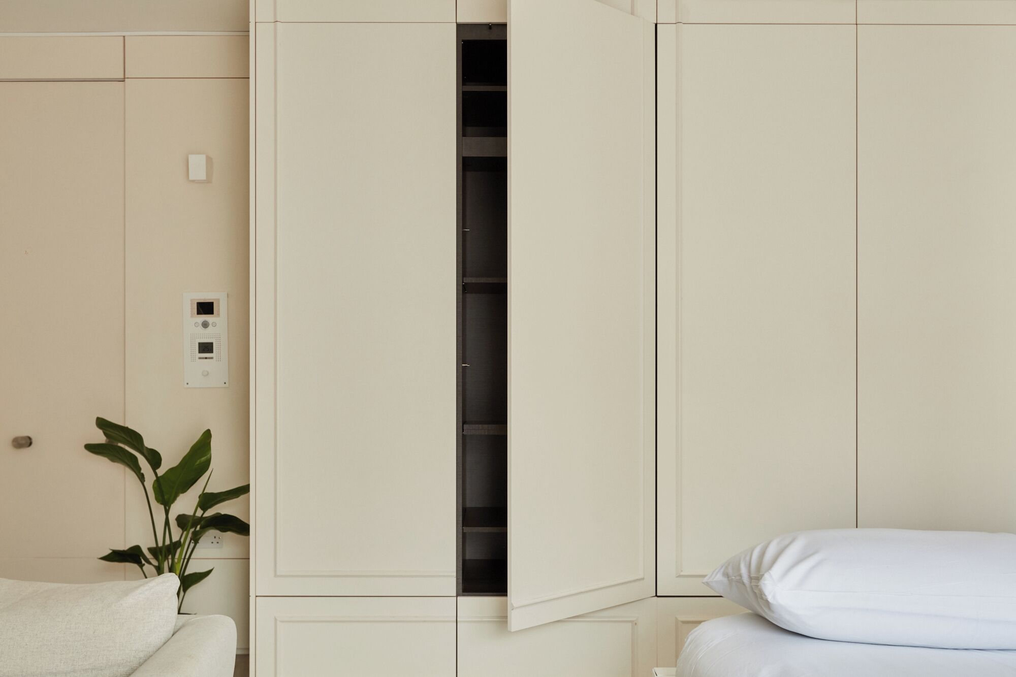 A bedroom with a bed, a white bedside table, a plant, and a wardrobe with a partially open door.