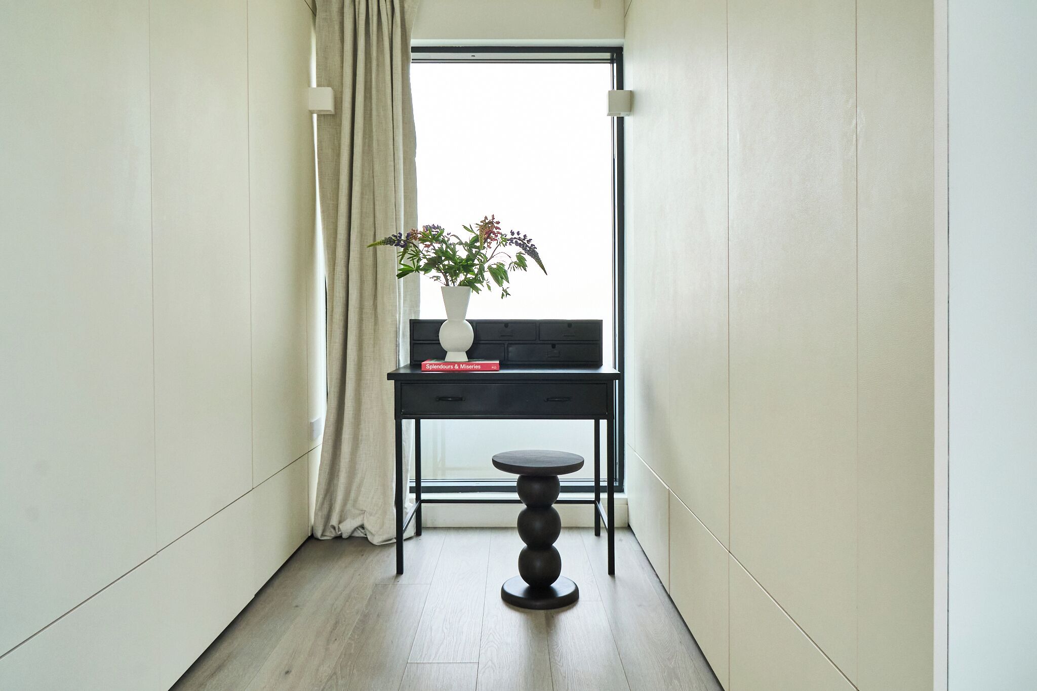 A narrow hallway with a desk, a vase with flowers, and a window with a curtain.