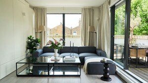 Un salon moderne avec une table basse en verre, un canapé gris et une vue sur un balcon aménagé avec du mobilier d’extérieur.