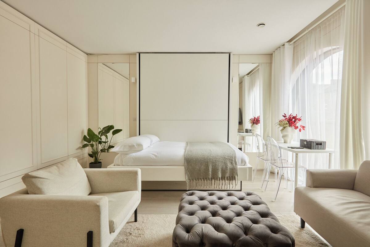 Ein modernes Schlafzimmer mit einem Bett, zwei Sofas, einem gepolsterten Hocker, einem kleinen Tisch und einer Zimmerpflanze.