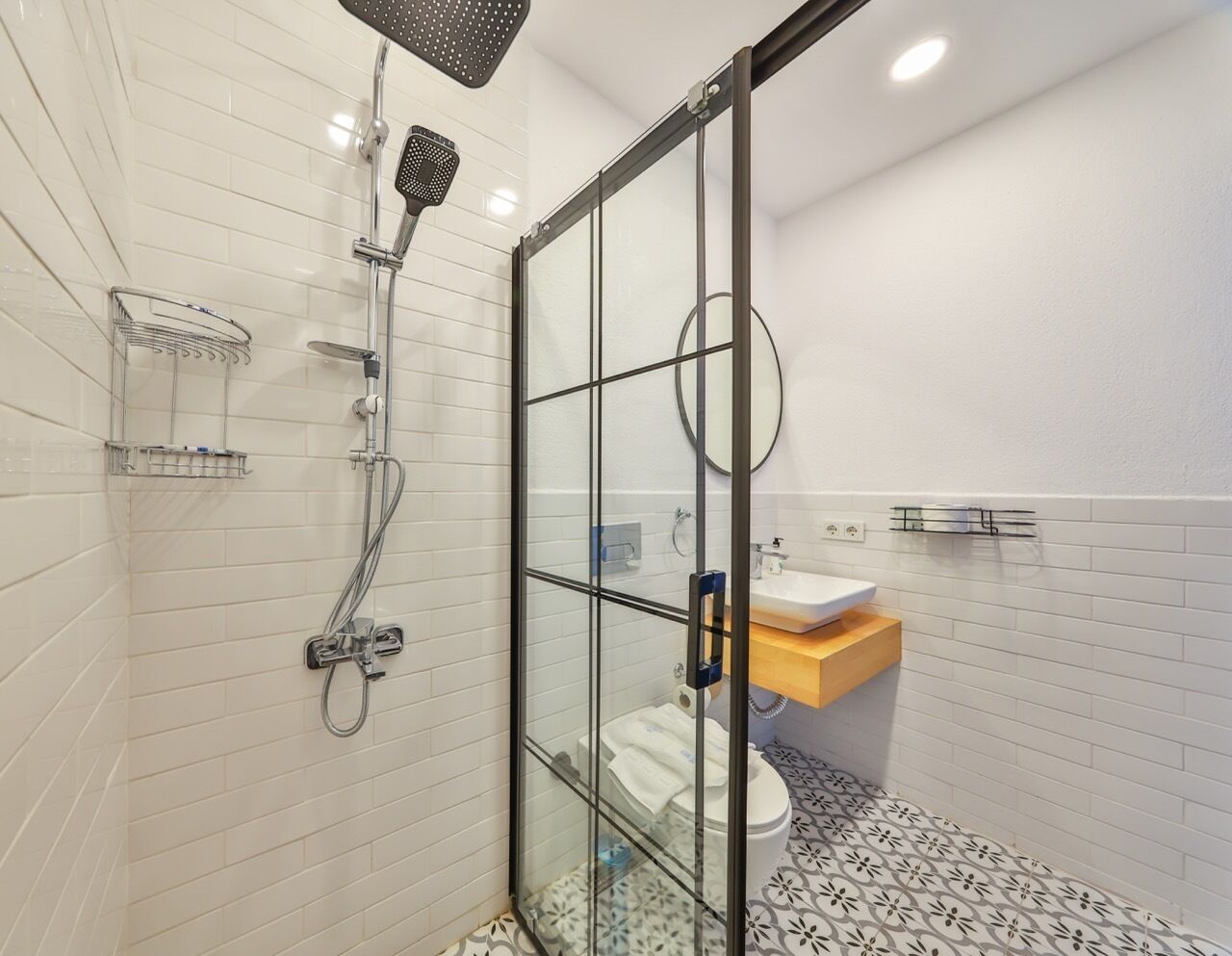 Une salle de bain moderne avec une douche à parois de verre, des murs recouverts de carreaux blancs, des toilettes et un lavabo doté d’un plan de travail en bois.