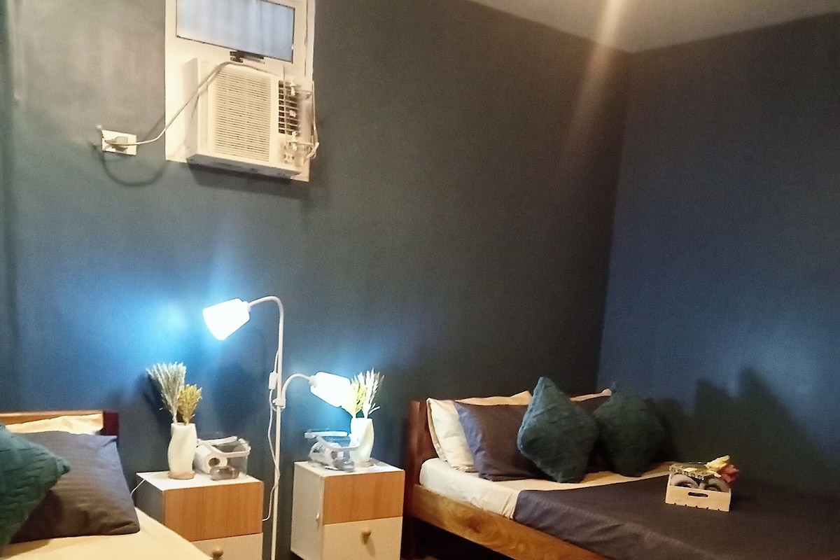 Una habitación con dos camas, un escritorio, una lámpara y una ventana con aire acondicionado.