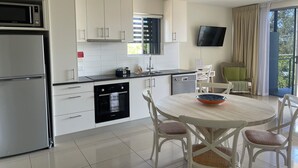 Una cocina moderna con armarios blancos, horno Bosch, microondas y lavavajillas. Hay una zona de comedor con una mesa redonda y sillas de mimbre. Un televisor de pantalla plana está montado en la pared, y se ve una ventana con cortinas.