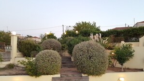 Un escalier en pierre mène à une entrée fermée par un portail, l’ensemble étant entouré d’arbustes et de petits arbres aux formes soignées.