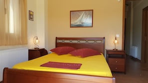 A hotel room with a wooden bed, yellow bedding, red pillows, two bedside lamps, a painting of a sailboat on the wall, and a radiator. - Villa Politimi at Aegina (Aegina)
