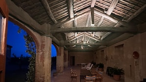 An outdoor patio with brick flooring, wooden tables and chairs, potted plants, and a rustic ceiling structure.