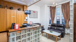 A reception area with a wooden reception desk, a television mounted on the wall, a sofa, and a window with curtains. - Abbey Hotel (London)