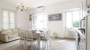 A bright, open-plan living and dining area with a sofa, dining table, chairs, a chandelier, and a kitchen with a stove.
