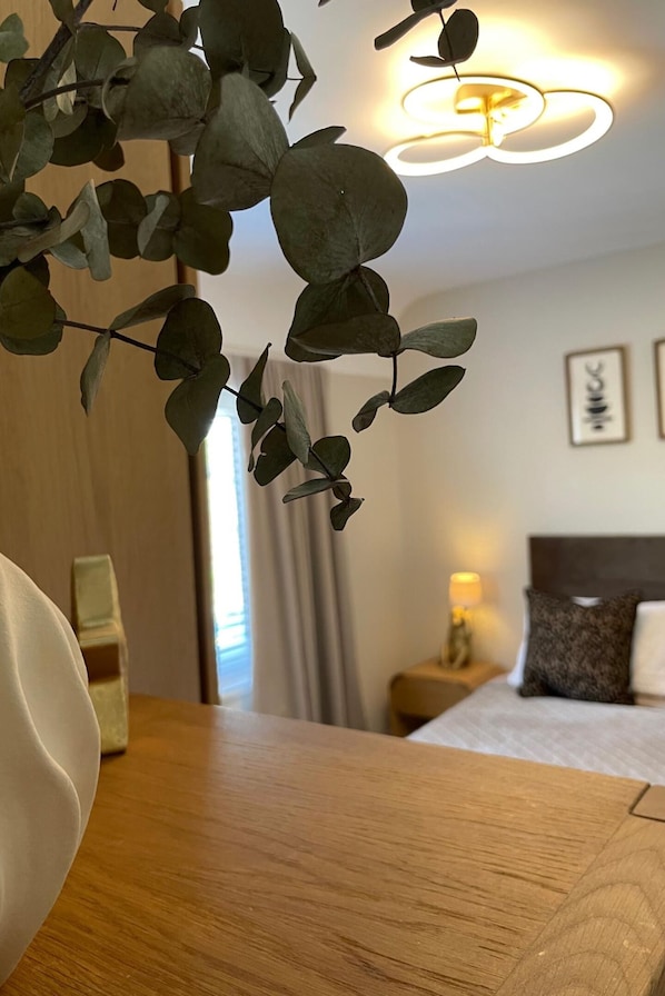 A hotel room with a wooden table, a bed with a headboard, a nightstand with a lamp, and a wall-mounted light fixture. - The Dooley, 3-beds, Sleeps 6, Pet Friendly (Bootle)