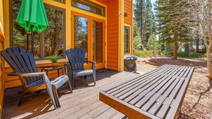A wooden deck with two Adirondack chairs and a green umbrella, a wooden table, and a house with yellow siding and large windows.