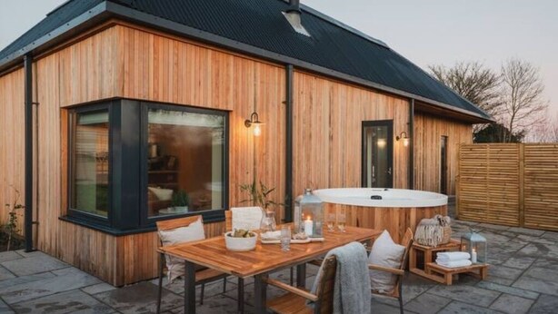 A wooden exterior building with a large window, a round outdoor table, and a seating area with cushions.