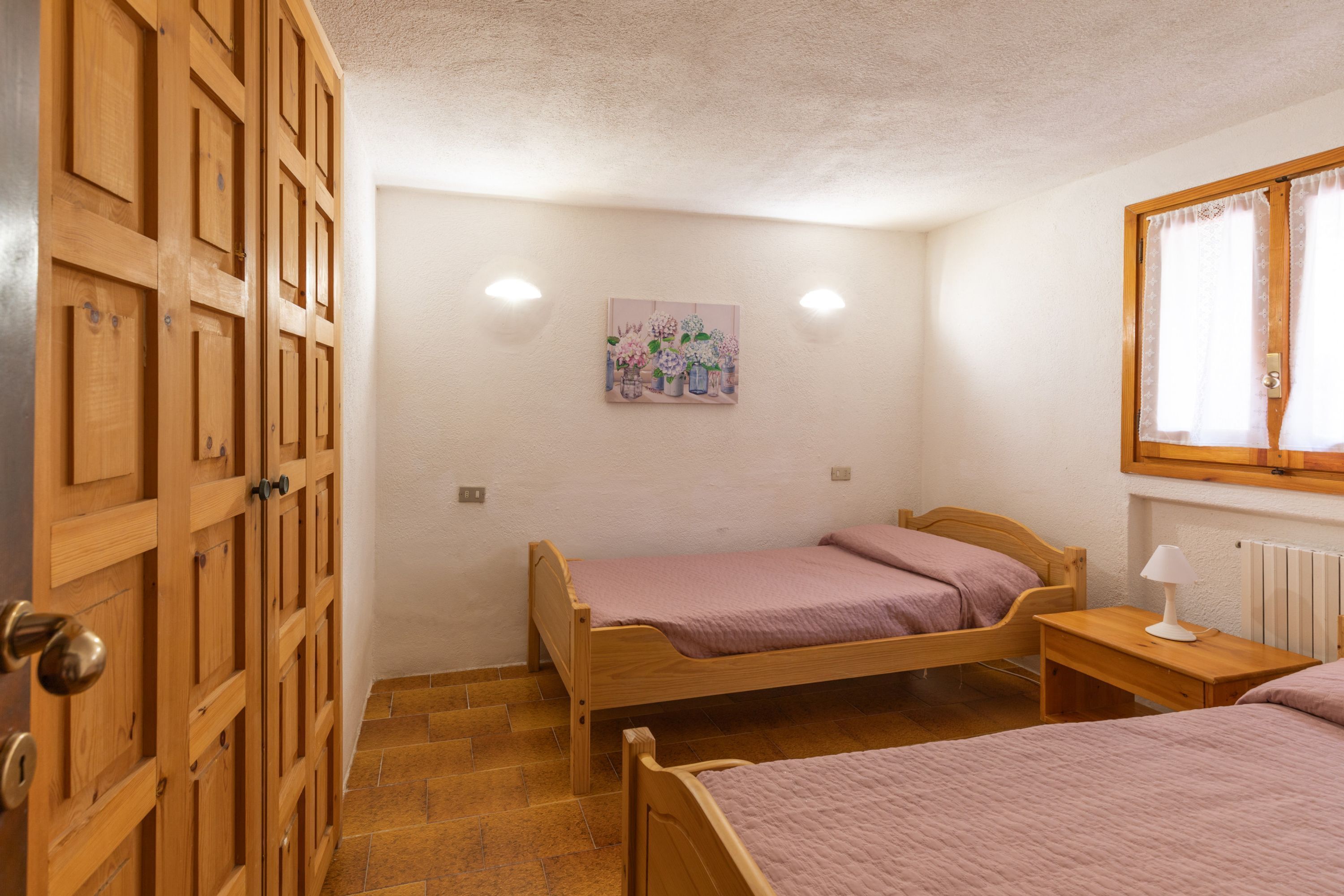 A bedroom with two wooden beds, a wooden wardrobe, a painting on the wall, and a window with curtains.
