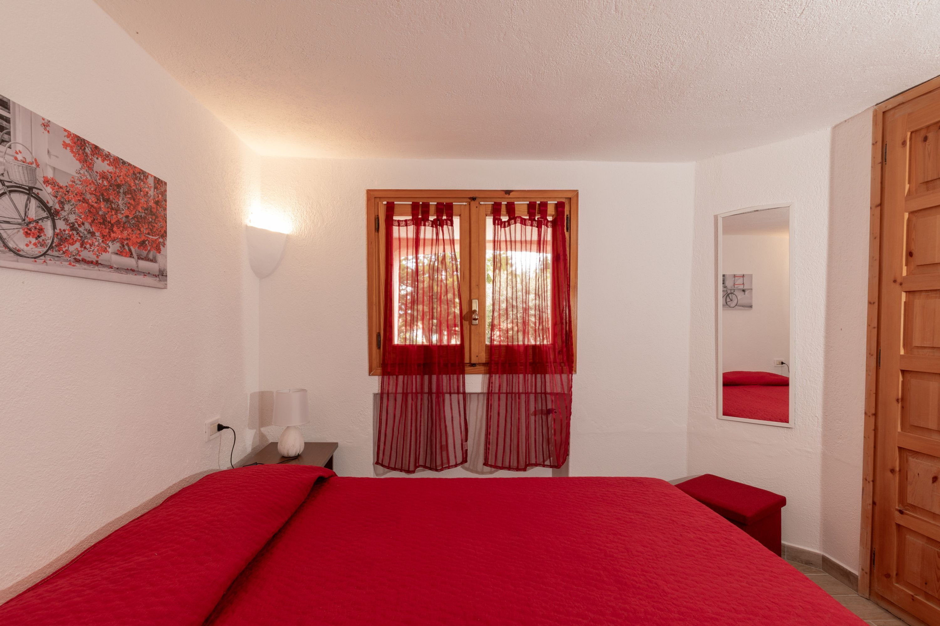 Ein Schlafzimmer mit roter Bettdecke, einem Fenster mit roten Vorhängen, einem Spiegel und einer Holztür.