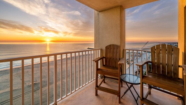 Un balcon avec une chaise en bois et une petite table, donnant sur une plage au coucher du soleil.