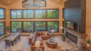 A spacious living room with a large fireplace, wooden floors, and large windows offering a view of the outdoors.