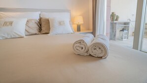 A neatly made bed with rolled towels, a nightstand with a lamp, and a view of the outside through a large window. - Dream Sardinia: Villa la Gioia, luxury with sea views and unique sunsets (Quartu Sant'Elena)