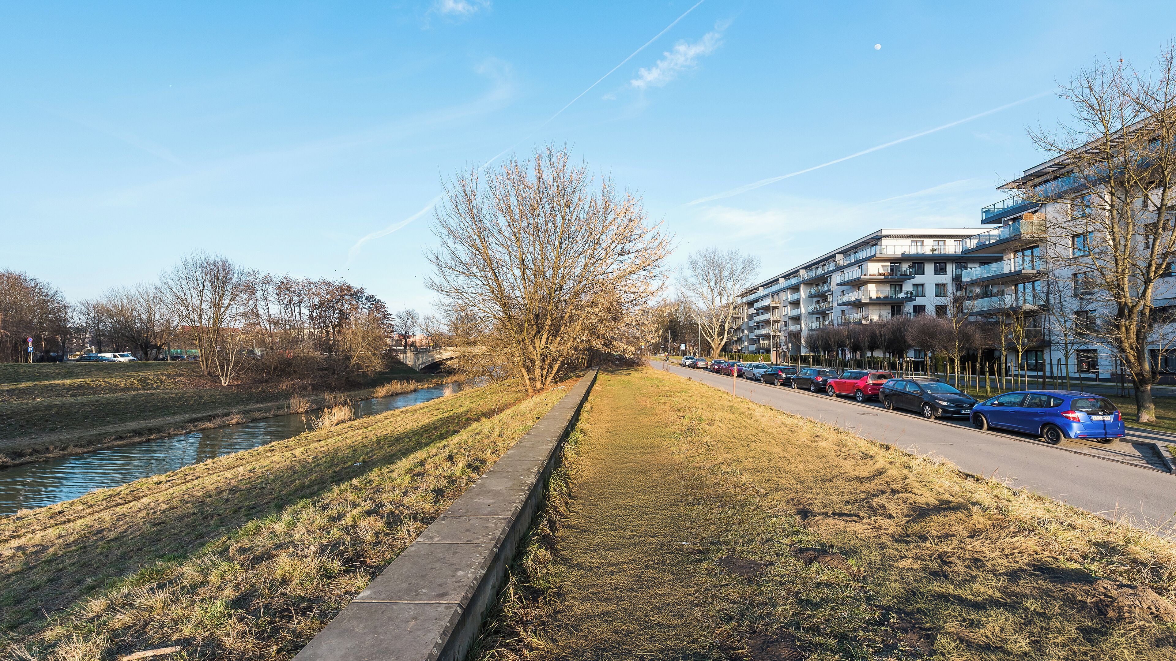 A riverside pathway with a concrete edge, a row of parked cars, and a modern apartment building with balconies.