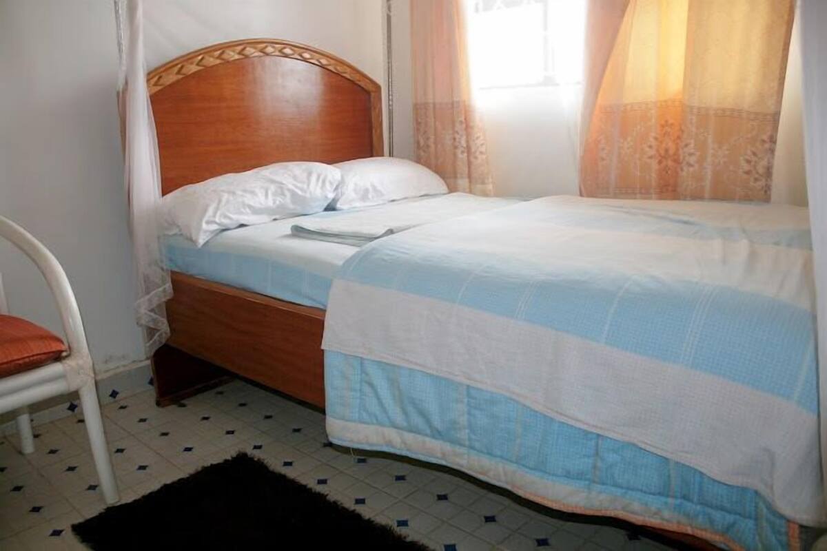 A single bed with a wooden headboard, white bedding, and a black rug on a tiled floor.