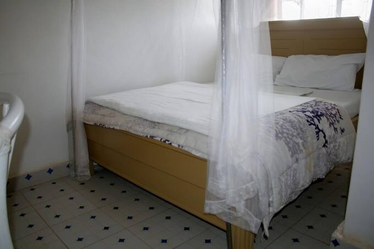 A single bed with a wooden headboard, a white bedspread, and a sheer canopy.