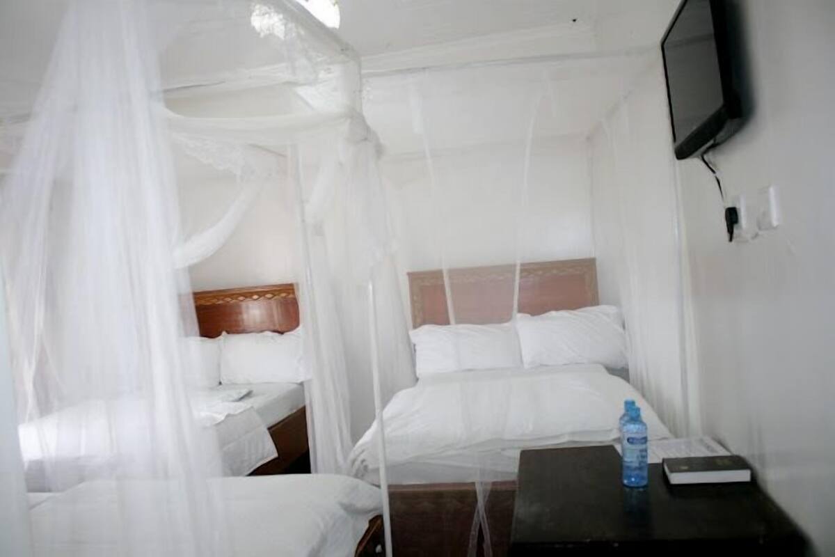 A room with two beds, one with a canopy and one without, a bedside table with a water bottle, and a TV mounted on the wall.