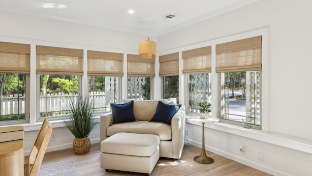 Ein geräumiges Wohnzimmer mit einem beigefarbenen Sofa, passendem Hocker und einem kleinen Beistelltisch. Große Fenster mit beigen Jalousien lassen viel natürliches Licht herein.
