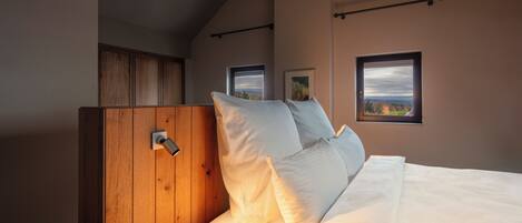 A bedroom with a wooden headboard, white bedding, and two windows with views.