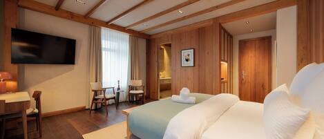 A modern hotel room with a wooden ceiling, a flat-screen TV, a desk with chairs, a large bed with white linens, and a bathroom visible through an open door.