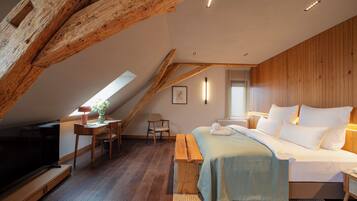 A bedroom with a sloped ceiling, wooden beams, a bed, a desk, a chair, a TV, and a window with blinds.