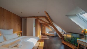 A modern bedroom with a sloped ceiling, wooden beams, a large bed, a flat-screen TV, and a view of the outdoors. - Hochblauen Hotel (Schliengen)