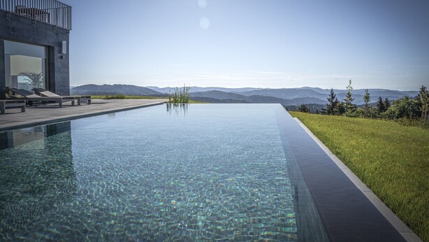 Una piscina infinita moderna con agua cristalina, un área de descanso con sillas y vistas a montañas distantes.
