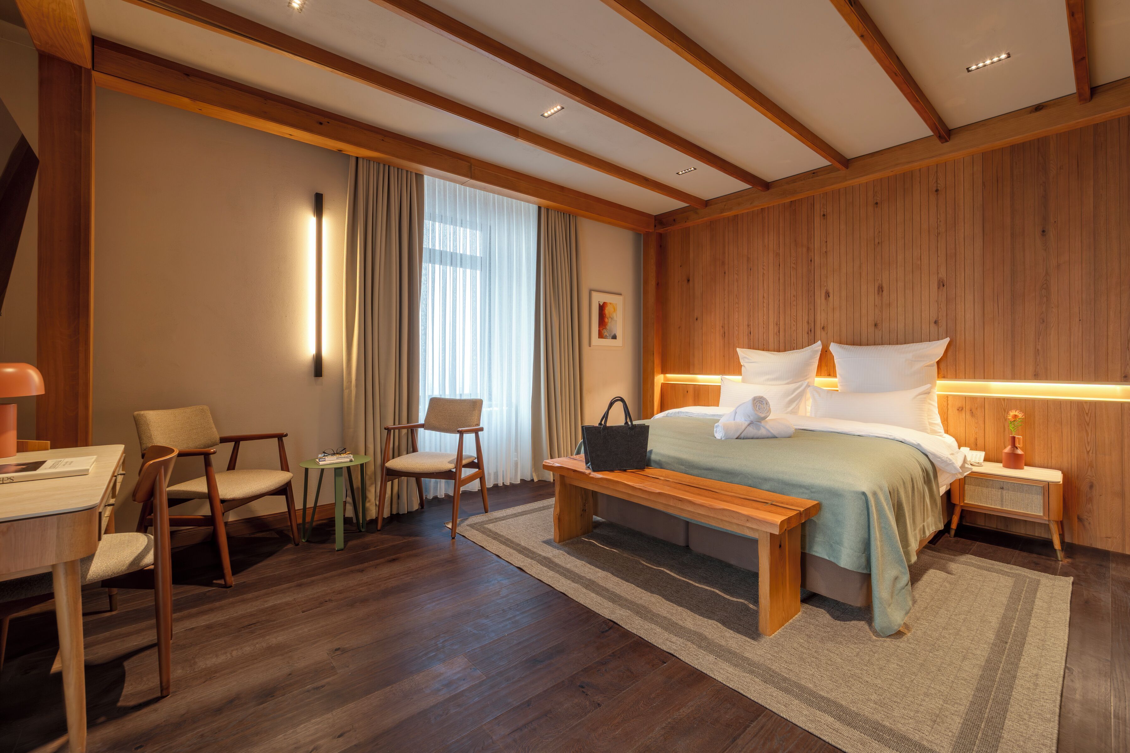 A modern hotel room with a wooden ceiling, a large bed with white linens, a desk with a chair, and a small table with a lamp.