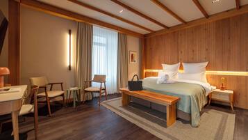 A modern hotel room with a wooden ceiling, a large bed with white linens, a desk with a chair, and a small table with a lamp.