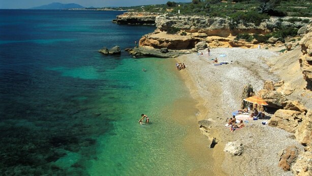 Una playa costera con agua turquesa cristalina, terreno rocoso y gente tomando el sol.