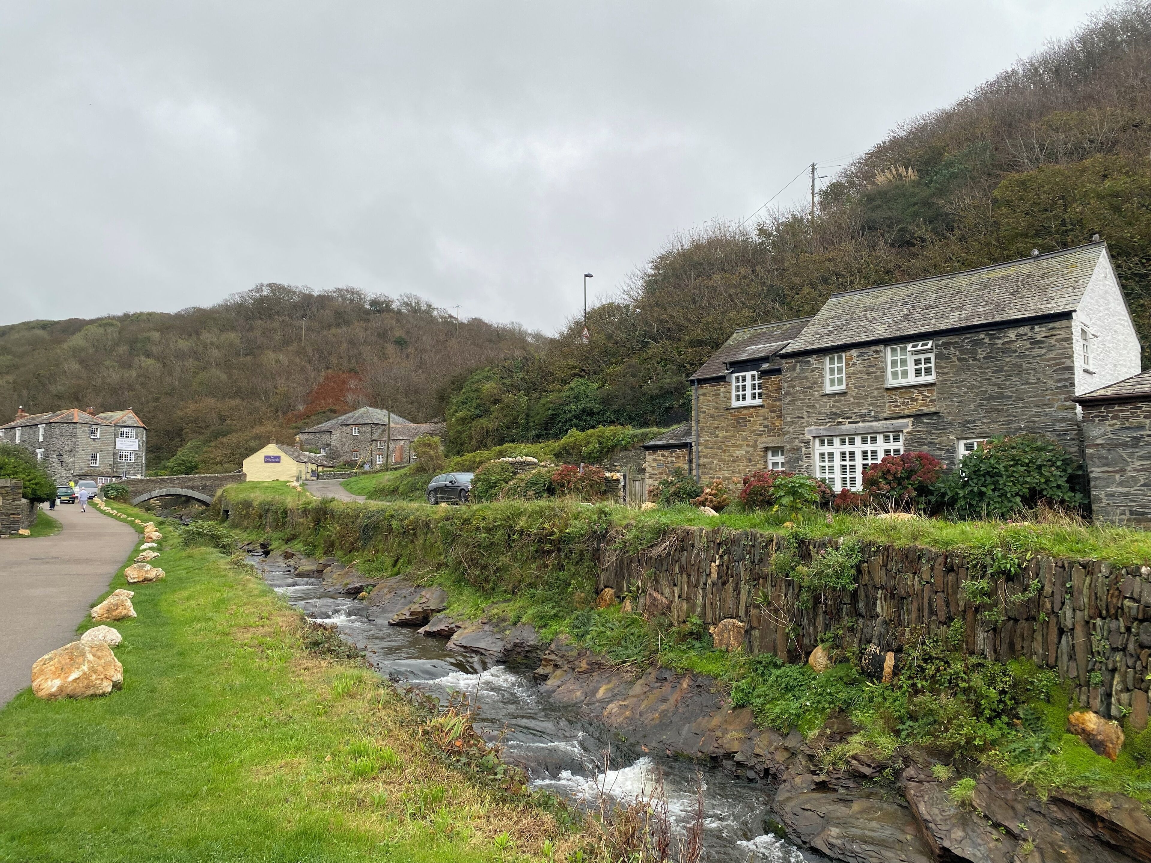 A stone building with a traditional stone roof, a grassy area, and a small stream running alongside a paved path.
