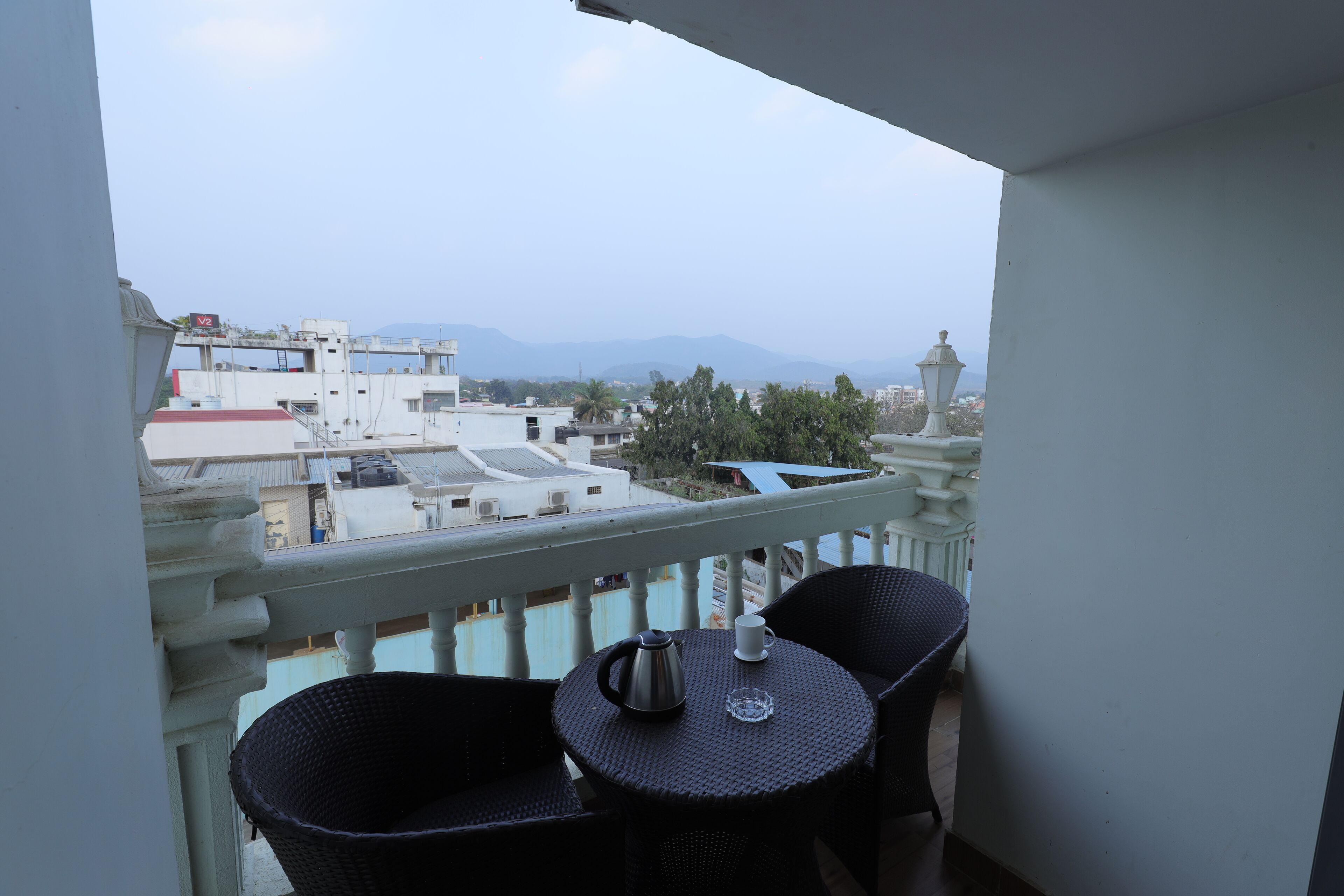 A balcony with a table and chairs, overlooking a cityscape with buildings and mountains.